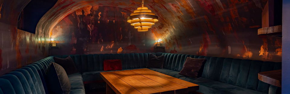 A dimly lit, barrel-vaulted room with teal velvet banquettes and a wooden table, illuminated by a tiered gold chandelier and wall sconces.