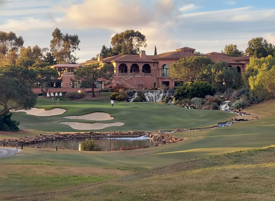 A large, ornate building with a red-tiled roof and multiple balconies overlooks a lush green golf course with sand traps and a pond, under a cloudy