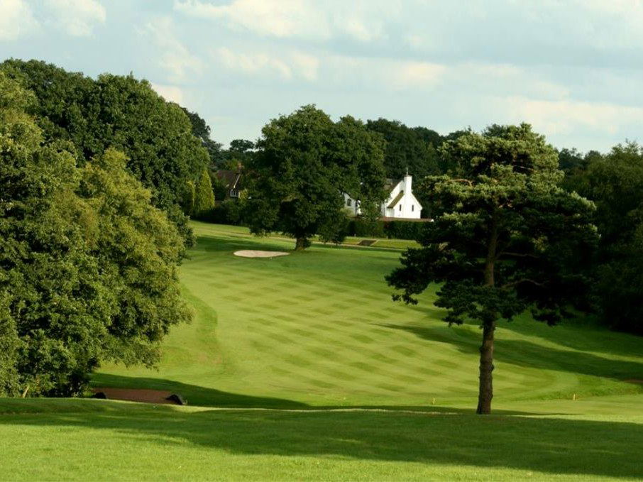 A wide-angle view of a well-maintained 18-hole championship golf course fairway lined with mature trees and a sand bunker, with a white clubhouse