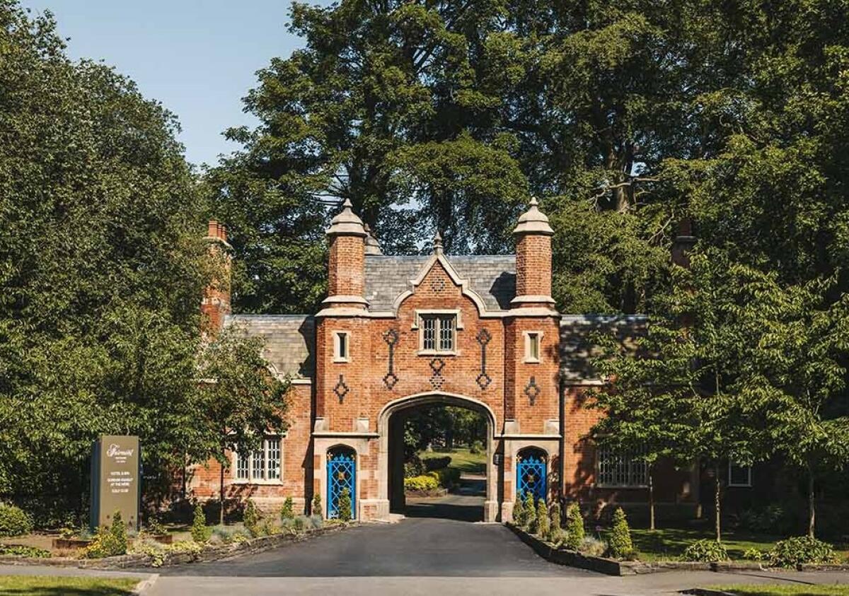 The entrance to Fairmont The Mere, a brick building with an archway and blue gates, surrounded by lush green trees.