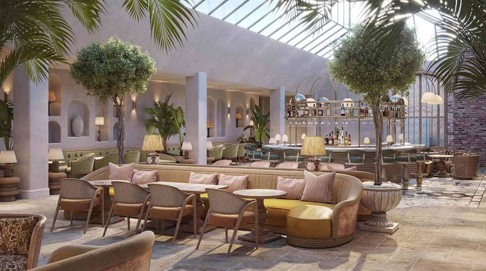 Rendered interior of The Orangery at Fairmont Cheshire, The Mere, showing a glass-roofed atrium with a curved marble bar, mature olive trees in stone urns, and wicker armchairs around curved banquettes.