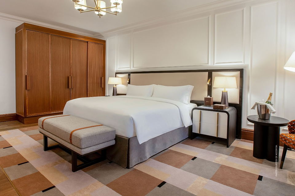 A hotel king bedroom with a large white-dressed bed, dark wood headboard, flanking table lamps, a tall timber wardrobe, geometric patterned rug, and a champagne bucket on a side table.