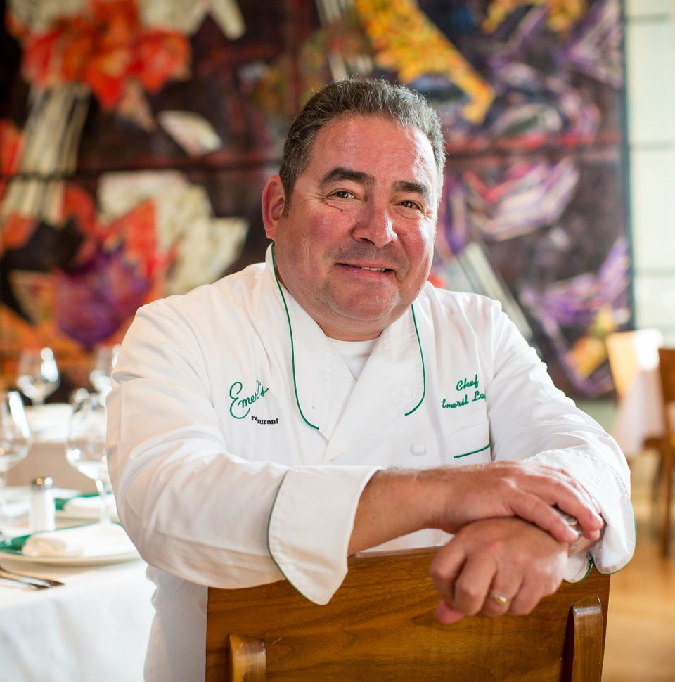 A chef in a white chef's coat with green trim and embroidery stands in a fine dining restaurant with colorful artwork visible in the background.