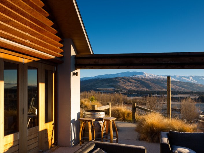 An outdoor patio with wooden barrel tables and stools, surrounded by golden grasses, overlooks a valley and snow-capped mountains under a clear blue sky. sky.