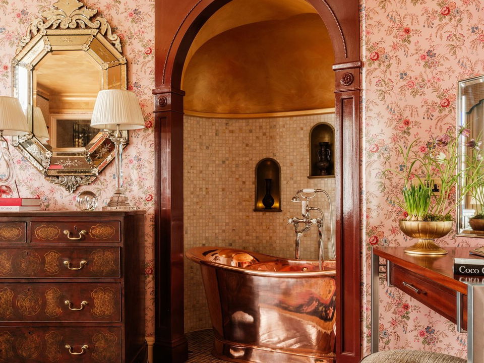 Copper freestanding bathtub set inside a dark-wood arched alcove with mosaic tile walls, surrounded by pink floral wallpaper, an ornate Venetian mirror, and a gilded chest of drawers.