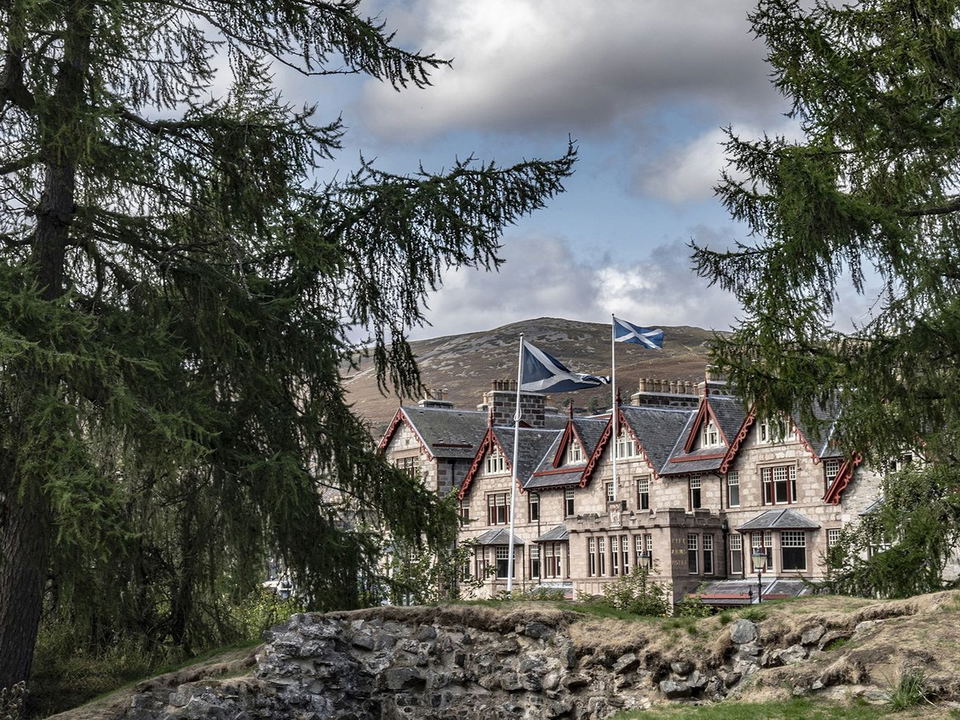 The Fife Arms hotel exterior, framed by conifers, with Scottish flags flying and a Highland hill behind.