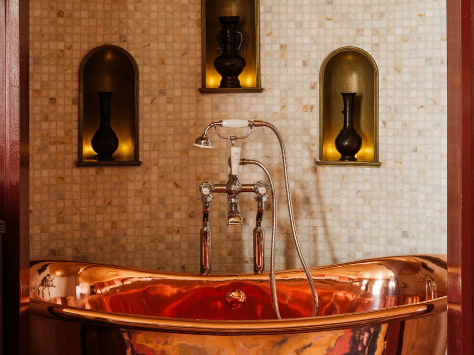A gleaming freestanding copper bathtub with chrome Victorian-style taps and hand shower, set against a mosaic-tiled wall with three arched alcove