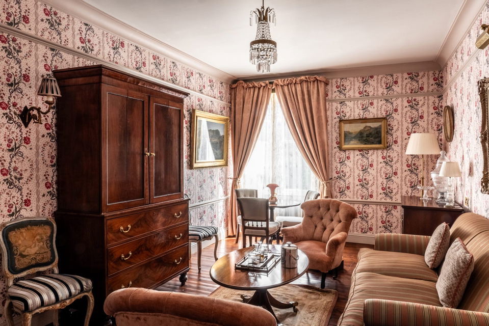 Antique-furnished sitting room with pink floral wallpaper, tufted velvet armchairs, mahogany armoire, crystal chandelier, and dusty pink drapes in the Coco Chanel Secret Room at The Fife Arms, Braemar.