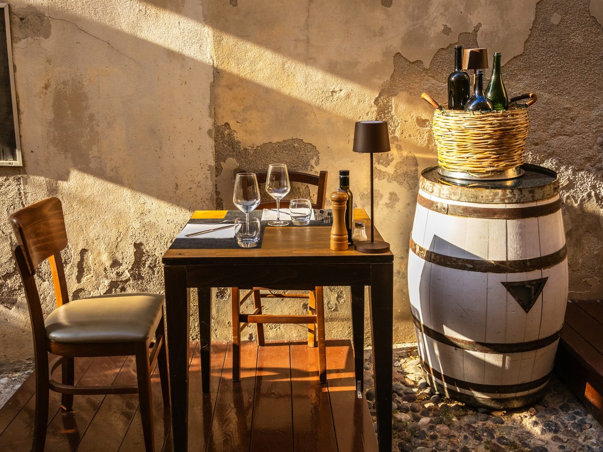 A rustic trattoria interior with a wooden table set for two, wine glasses, and a wicker basket of bottles on a barrel.
