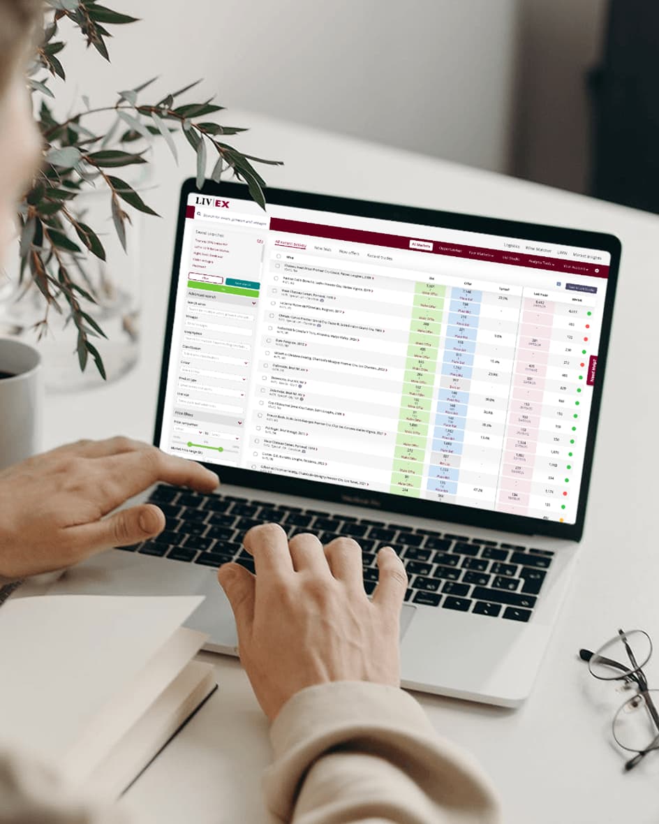 A person's hands typing on a laptop displaying the Liv-ex fine wine trading platform, showing market data and trade information.