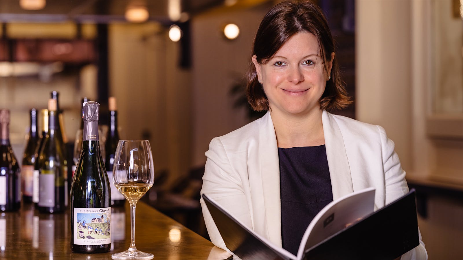 A woman in a white blazer and dark top smiles while holding an open book, with a bottle of Champagne Chavot and a glass of wine on a bar.