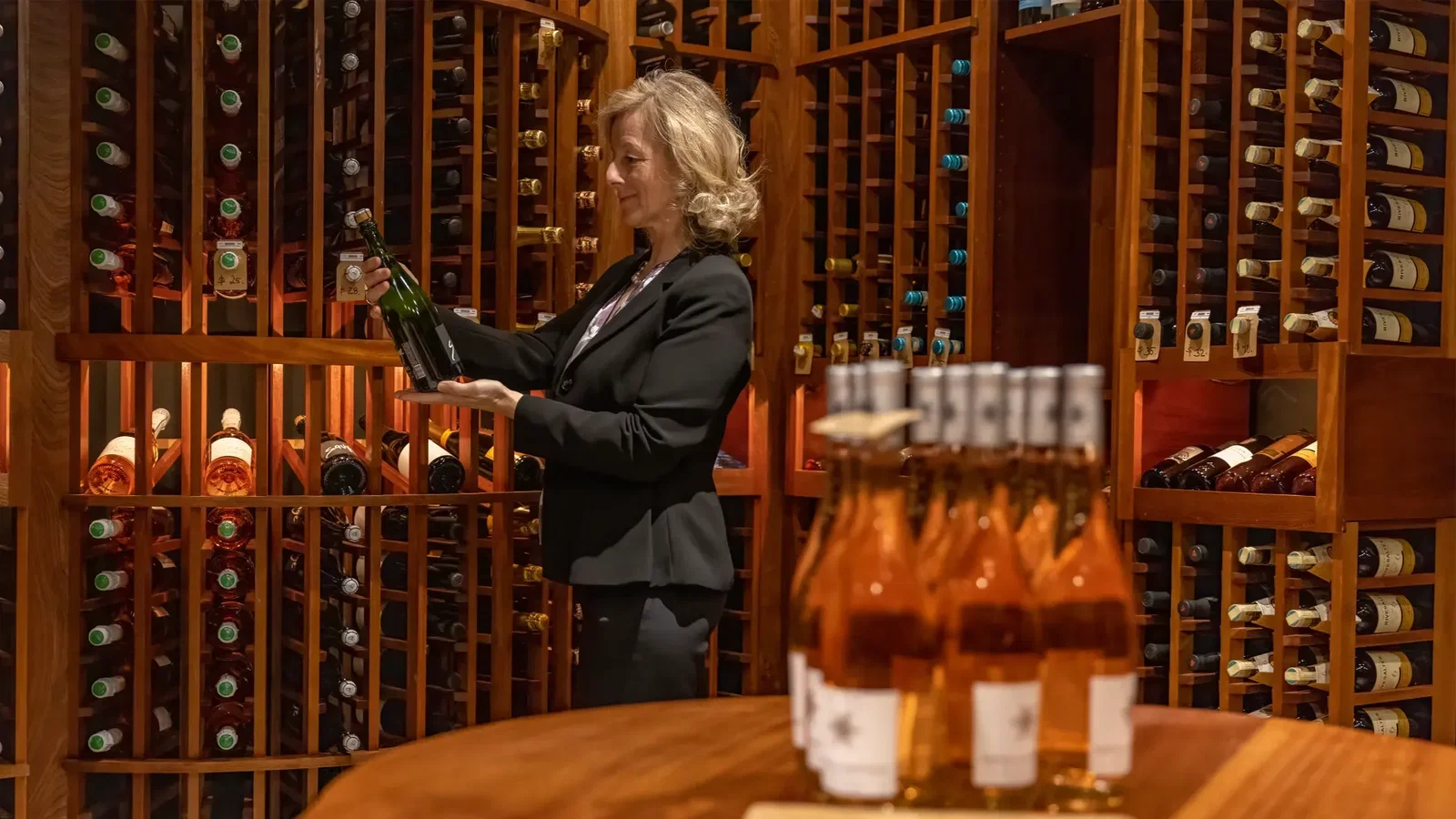 A woman in a black blazer holds a green wine bottle, examining it in a dimly lit wine cellar with wooden racks.