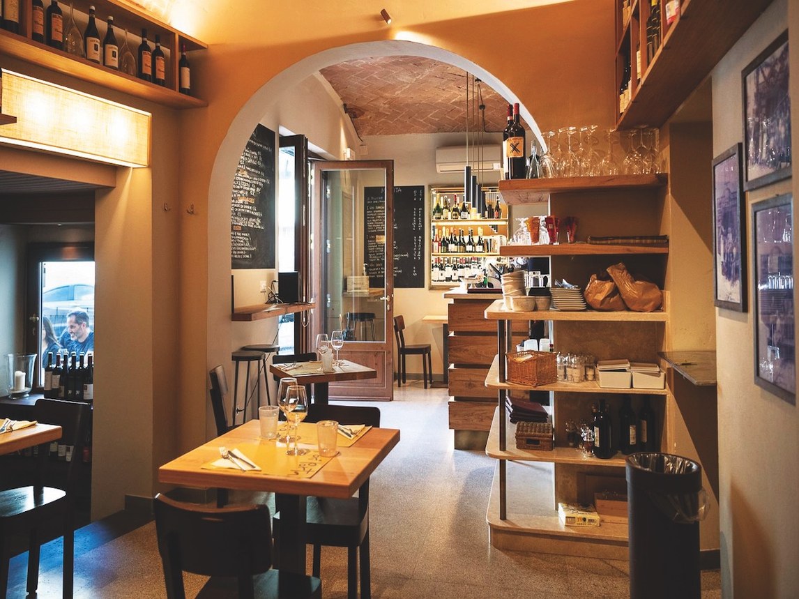Interior dining room of a rustic Italian restaurant with warm ochre walls, arched brick ceiling, wooden tables set with wine glasses, open shelving wi