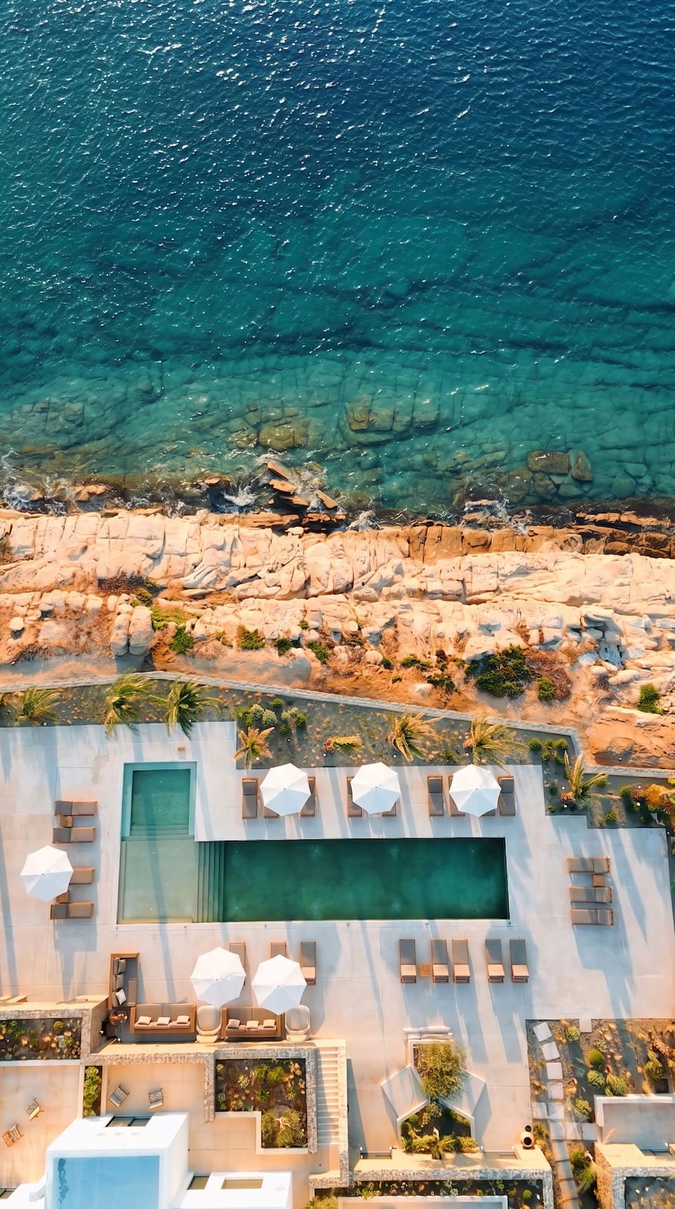 Overhead shot of a modern white resort building with a central pool, sun umbrellas, and loungers overlooking a pristine Mediterranean beach with turquoise water and rocky shores.