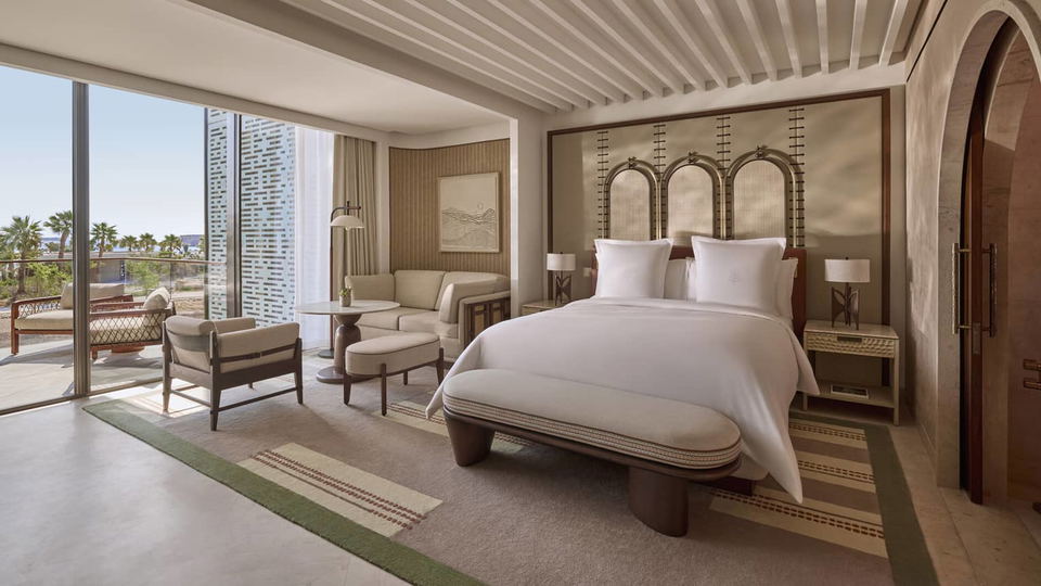 A spacious hotel room with a large bed, a sitting area with a sofa and chairs, and a balcony overlooking palm trees and the sea.