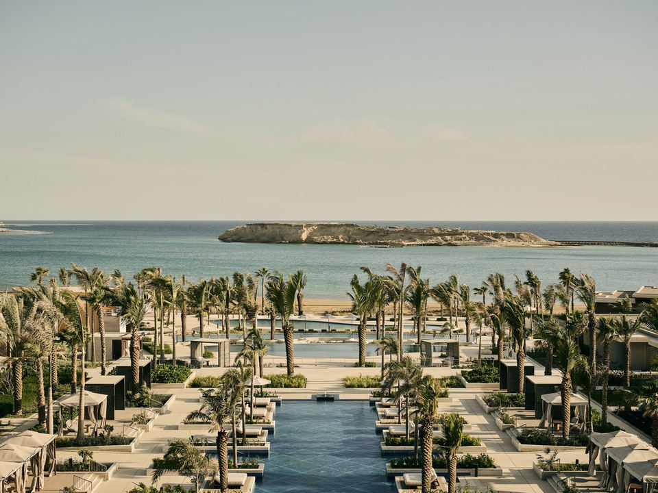 An aerial view of the Four Seasons AMAALA Triple Bay resort, featuring symmetrical pools, palm trees, cabanas, and the Red Sea coastline.