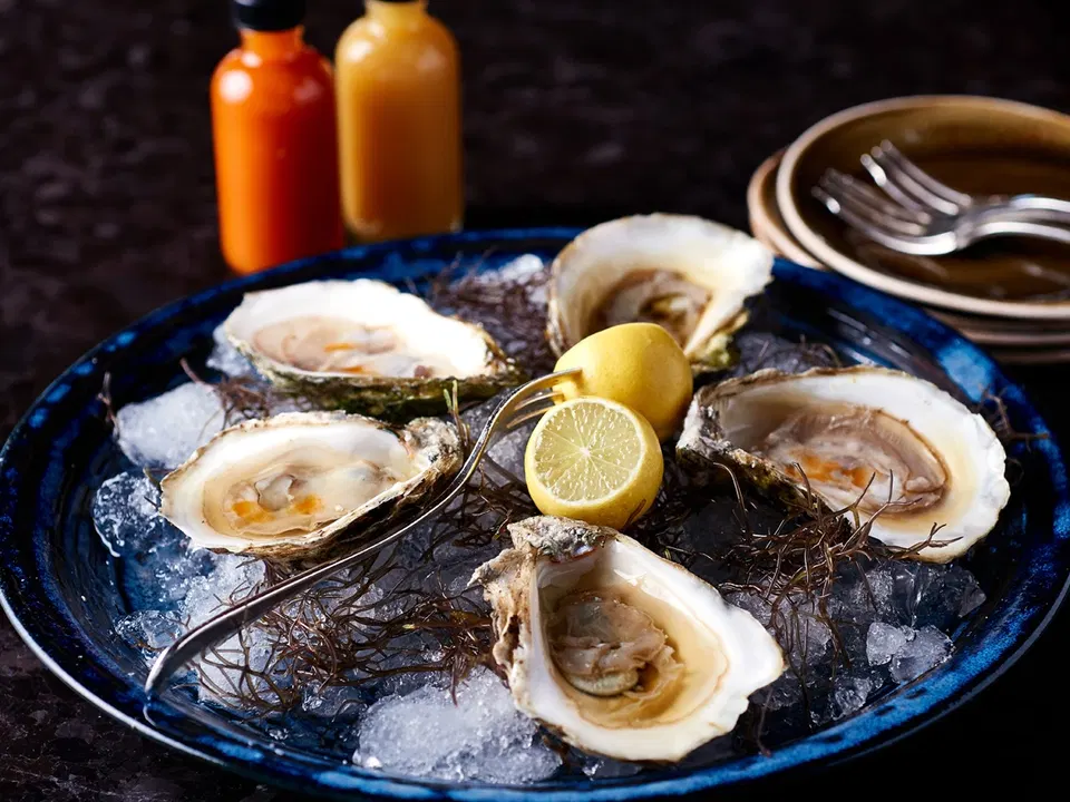 A blue plate of fresh oysters on ice, garnished with seaweed and lemon halves, with two bottles of hot sauce and stacked plates with forks in the