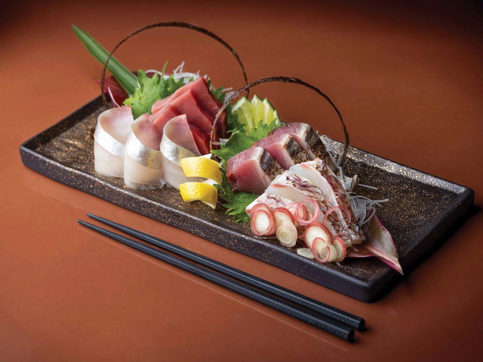 A beautifully plated sashimi omakase spread on a dark rectangular tray with chopsticks, featuring white fish, tuna, seared fish, and octopus/squid