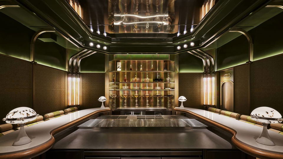 A modern bar with a mirrored ceiling, sleek metal counter, and shelves displaying various bottles and glasses, illuminated by warm lighting.