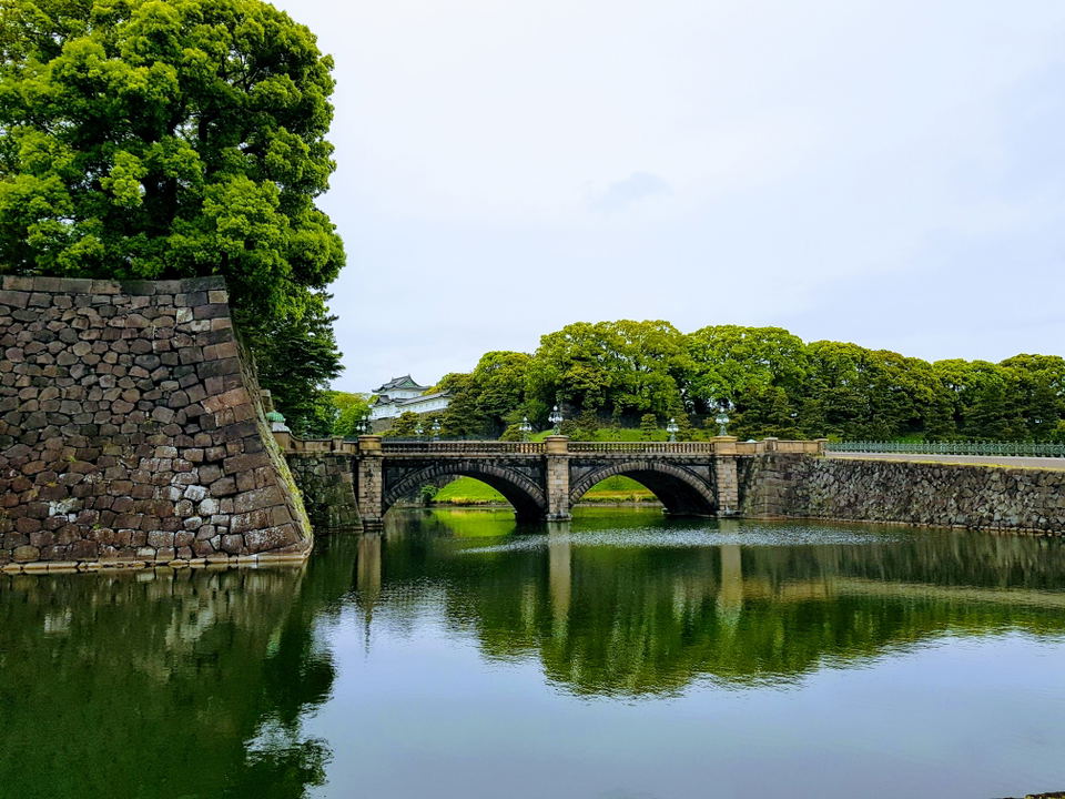 A stone bridge with multiple arches spans a green moat, leading to a traditional Japanese building with a dark roof nestled among lush green trees.