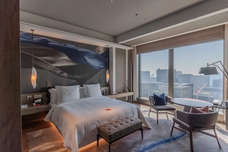 A modern hotel room with a large bed, two armchairs, a small round table, and a floor-to-ceiling window overlooking a city skyline.
