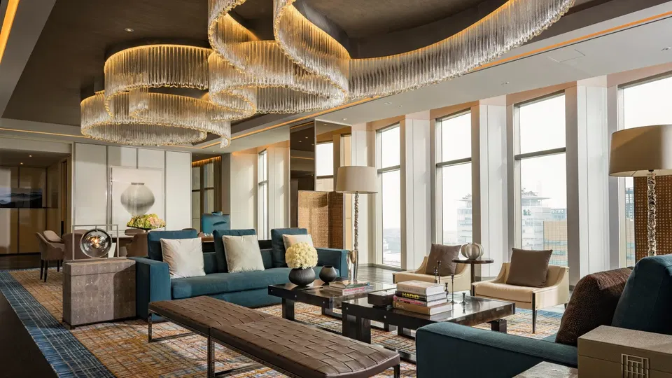 A luxurious living room with a large, ornate chandelier, teal sofas, and floor-to-ceiling windows offering city views.