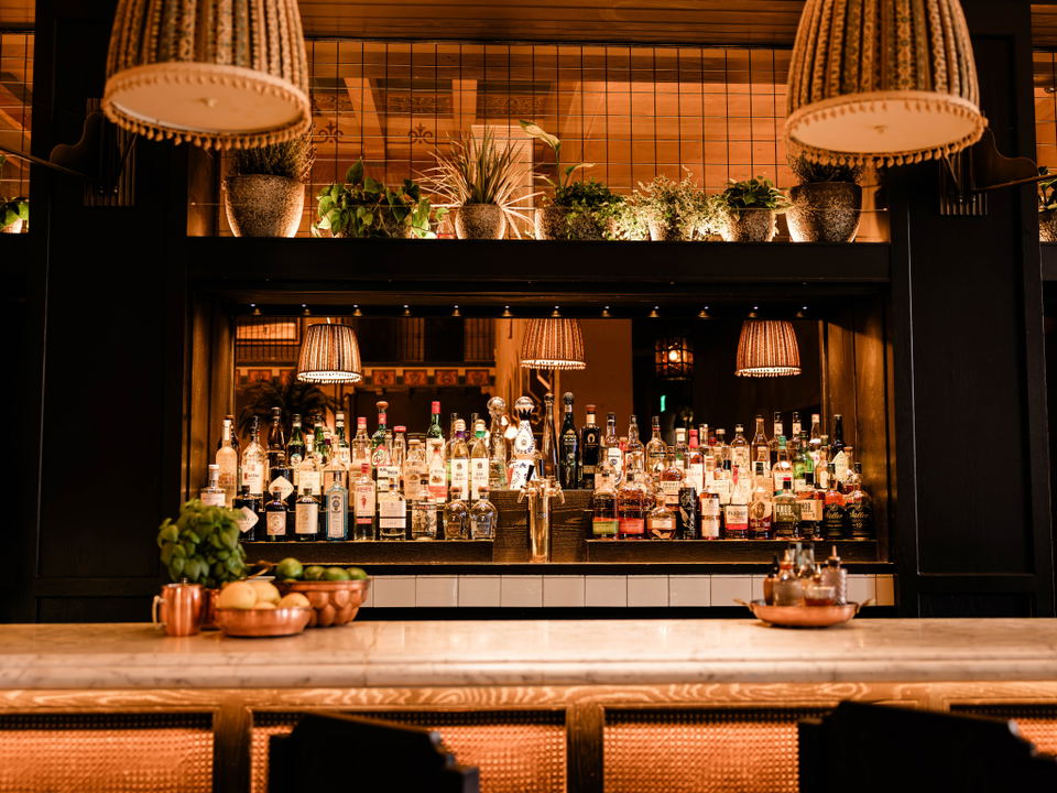 A well-lit bar interior with numerous bottles of spirits arranged on shelves, hanging woven pendant lights, wooden counter seating, and potted plants on the upper level.