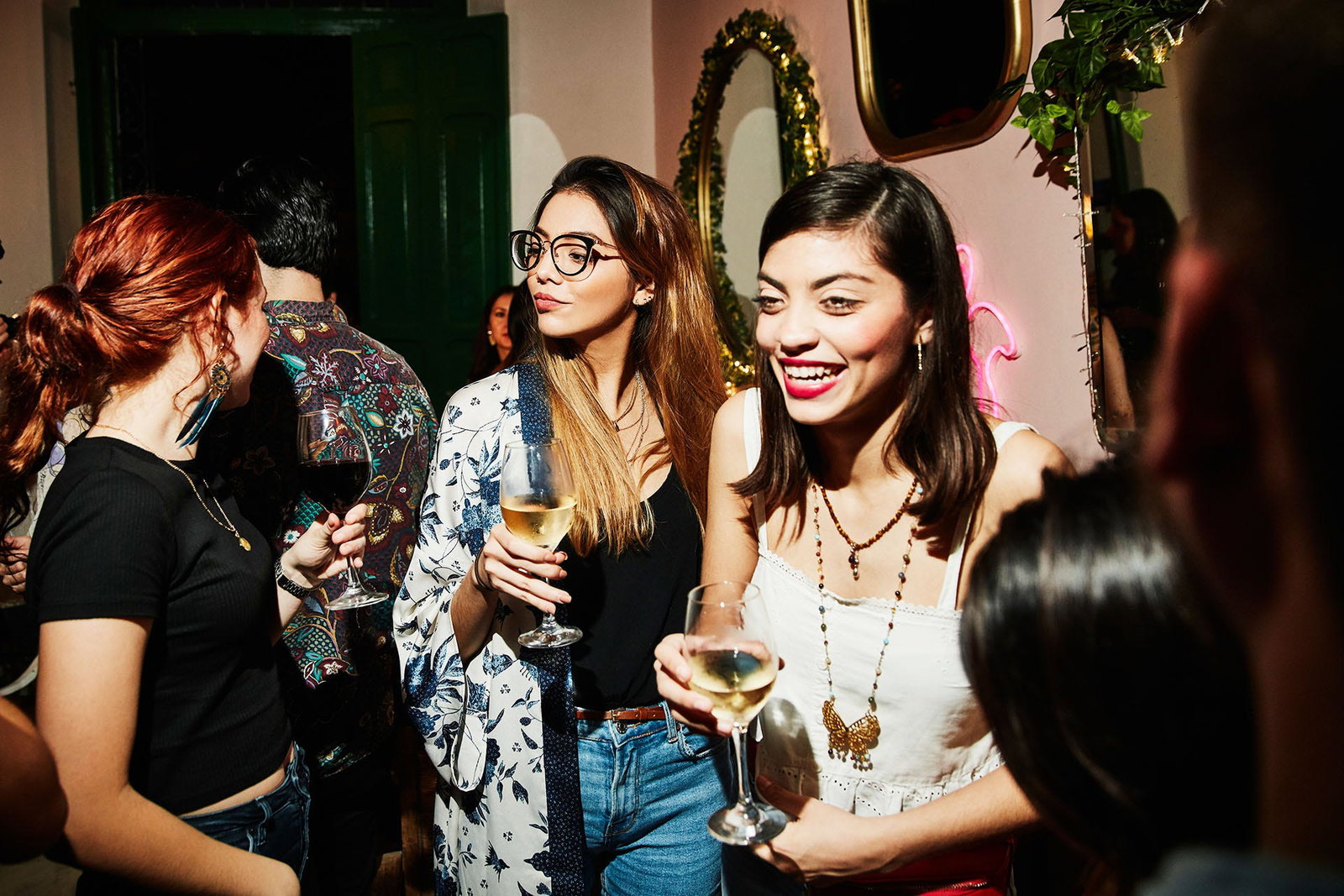 Two young women holding white wine glasses laugh and mingle at a lively, dimly lit bar alongside friends.
