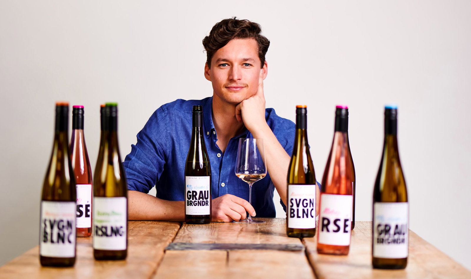 Young man in blue shirt resting chin on hand, seated at a wooden table surrounded by eight Young Poets wine bottles and an empty wine glass.