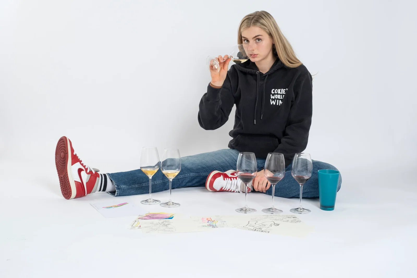 Blonde young woman in a black "Cokie's World of Wine" hoodie, jeans, and red Air Jordan sneakers sits on a white floor, sipping white wine from a glass surrounded by red wine glasses and sketches.