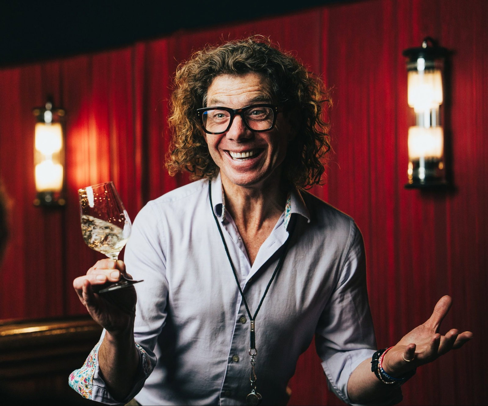 Curly-haired man with black glasses and a wide smile holds a white wine glass in a red-curtained room with wall sconces.