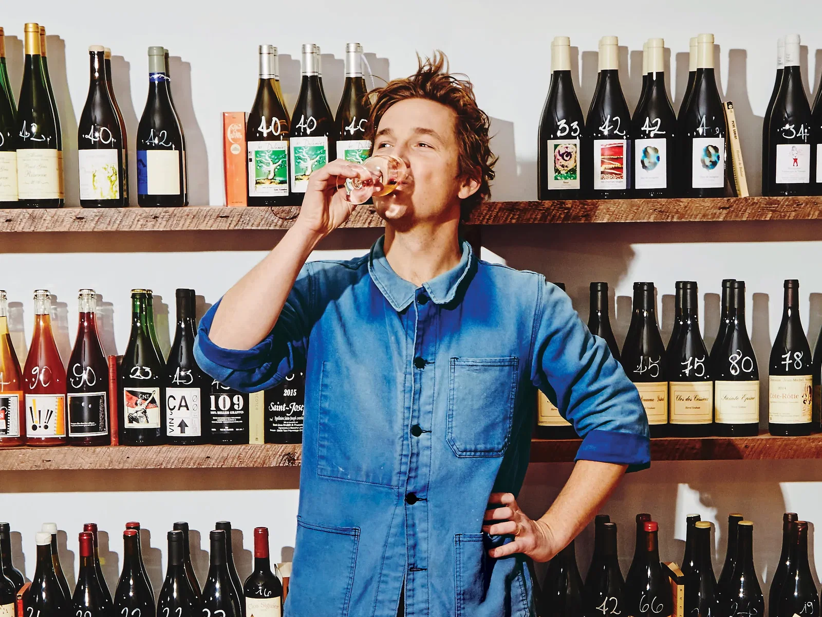 Bradford Taylor in a blue denim jacket sips wine in front of wooden shelves lined with natural wine bottles marked with handwritten prices.