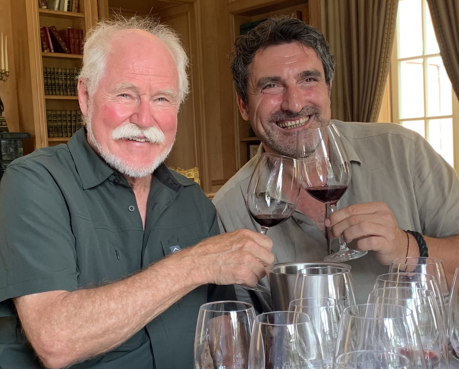 Two smiling men hold glasses of red wine at a tasting table, the man on the left with a white mustache and the man on the right with dark hair identified as winemaker Patrick Honnef.