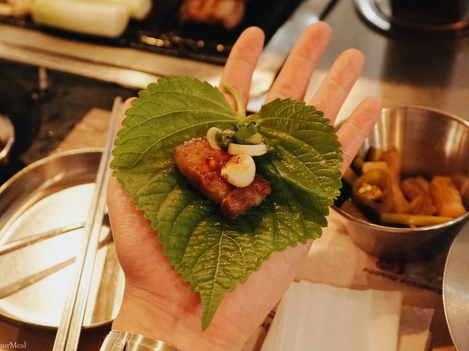A hand holds a perilla leaf wrap with a piece of grilled pork, sliced green onion, and garlic, consistent with Geumdwaeji Sikdang's Snow Flower Pork