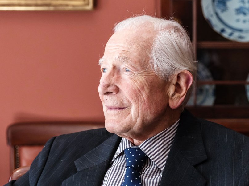 Elderly man in a dark pinstripe suit and navy polka-dot tie seated in a leather chair, gazing to his left, with a blue-and-white porcelain plate on shelves behind him.