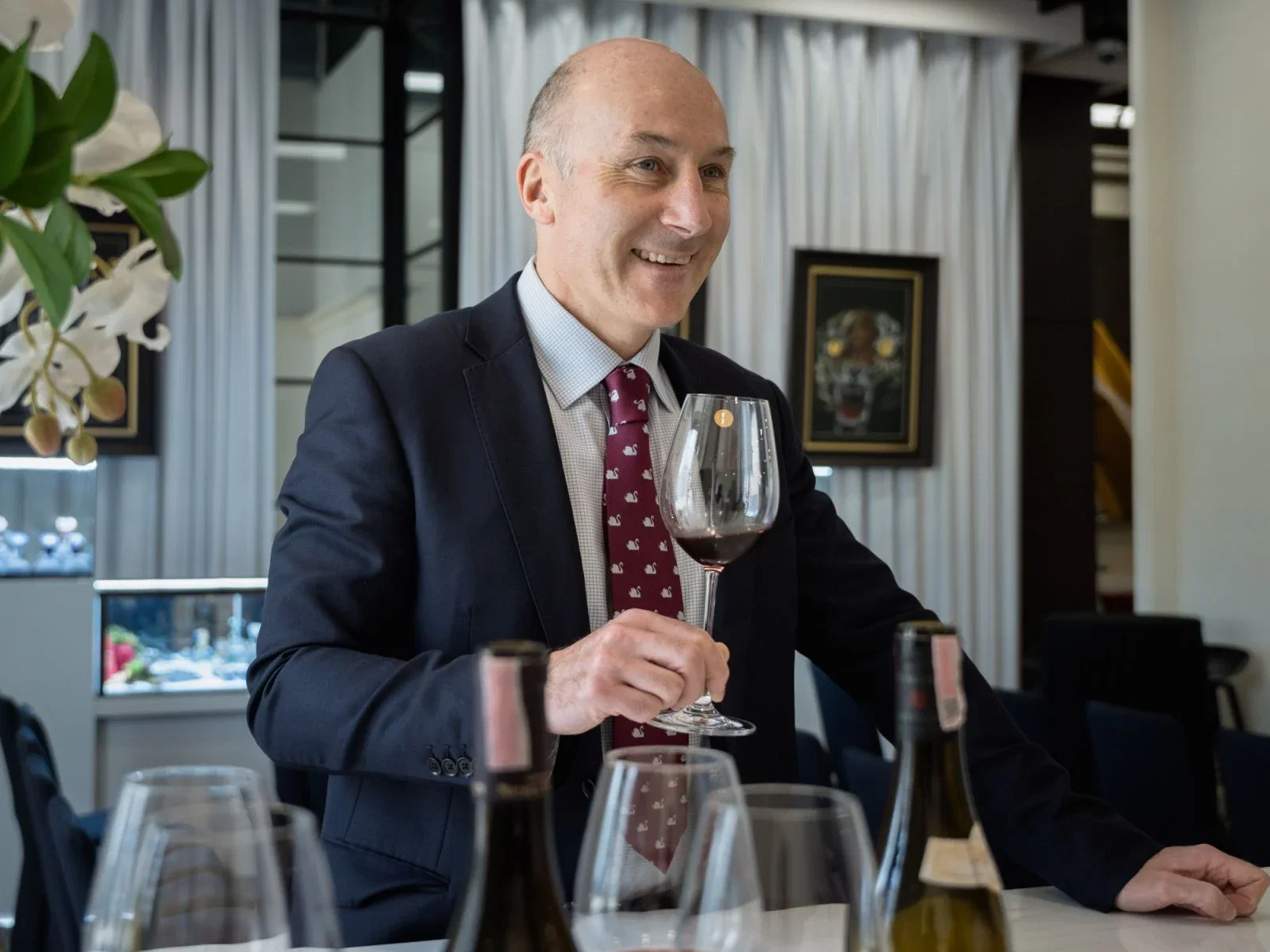 Tim Triptree, a bald man in a suit and tie, smiles while holding a glass of red wine, with other wine bottles and glasses on a table in front of him.