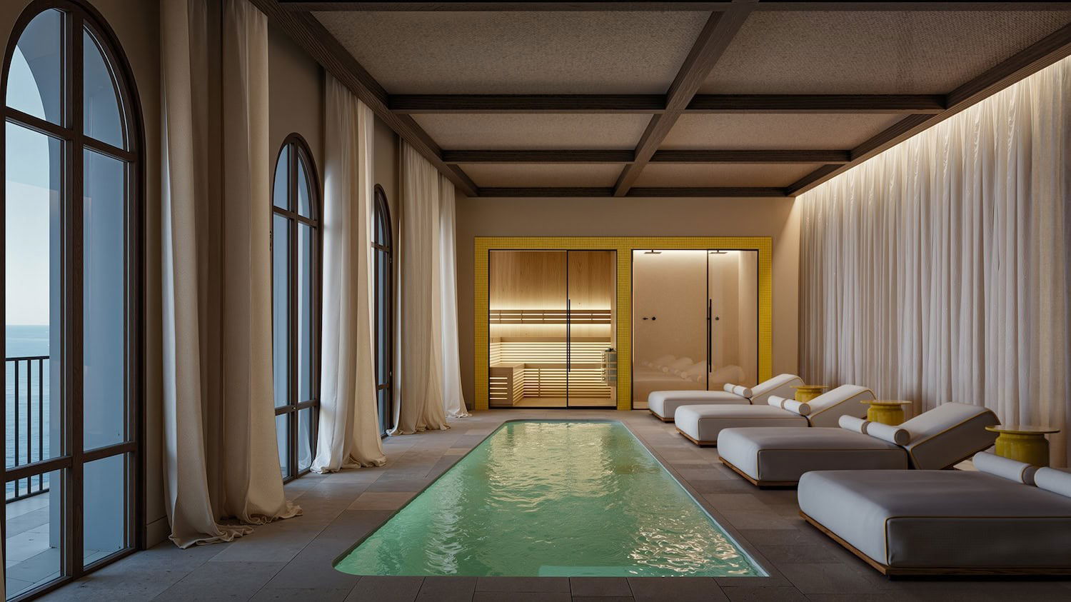 A luxury spa interior with a rectangular pool, white loungers, a sauna, and arched windows looking out to the ocean.