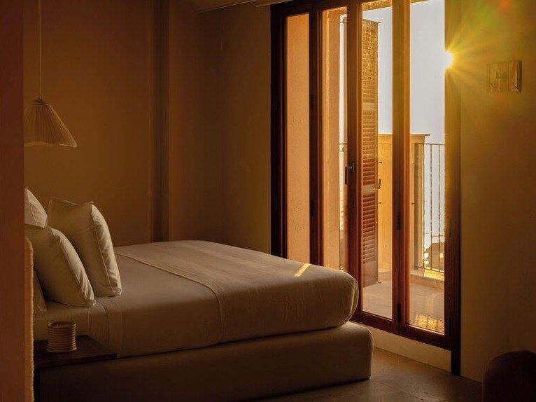 A hotel guest room interior at Gran Hotel Margalida with sun-washed yellow walls, white bedding, and French doors to a balcony.
