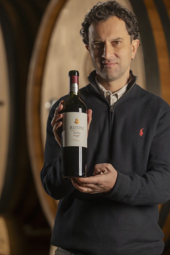 A man with curly hair in a dark blue sweater holds a bottle of Rutini wine with both hands, standing in a cellar with large wooden barrels in the