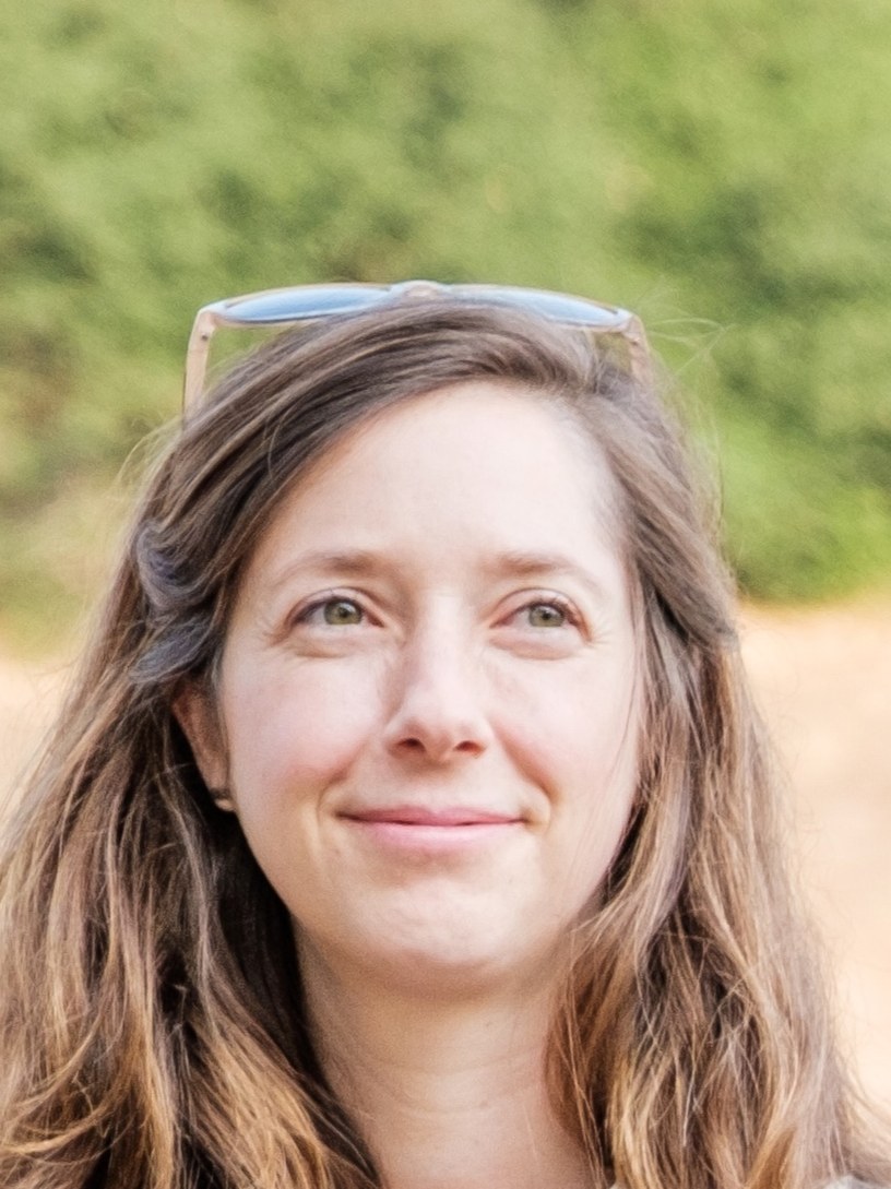 Hailey Trefethen, with long brown hair and sunglasses on her head, smiles in an outdoor vineyard setting.