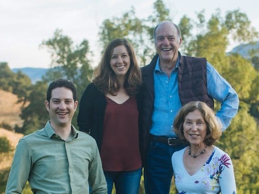 Four people and a golden retriever pose outdoors in front of a stone wall, with rolling hills and vineyards in the background.