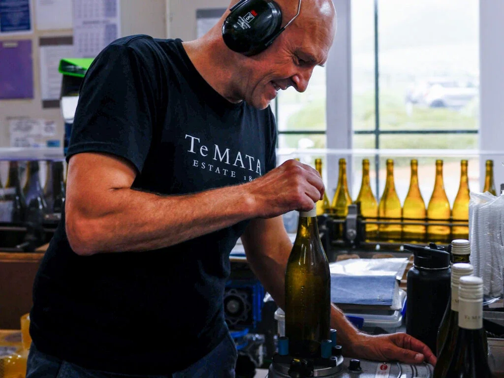A man wearing a black 'Te Mata Estate' t-shirt and ear protection works with a wine bottle on a workbench in a winery setting, with rows of green