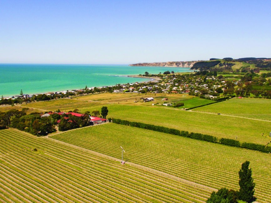 Aerial view of a coastal town with vineyards and green fields in the foreground, and the ocean and cliffs in the background under a clear sky.