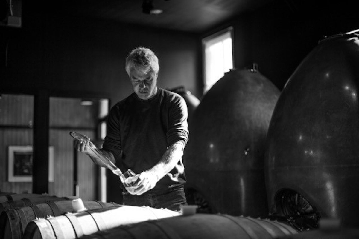 Winemaker Tony Bish using a wine thief to draw a barrel sample in a dimly lit winery, with large concrete egg fermenters beside him.
