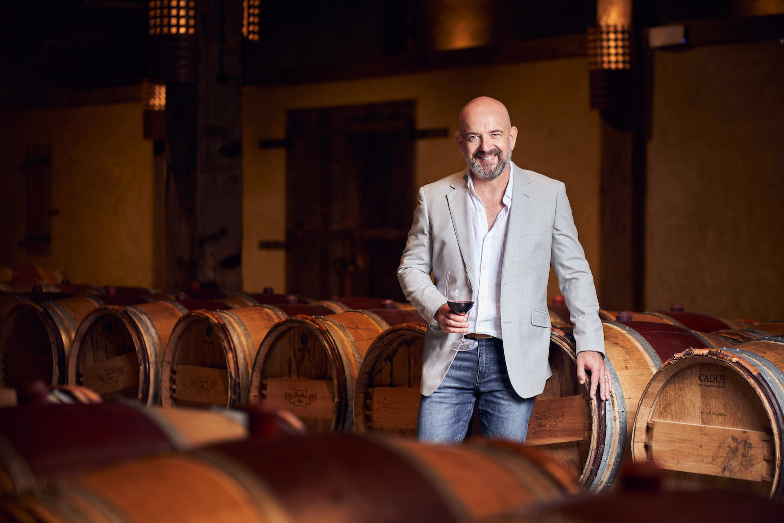 Bald man with beard in a light blazer holds a glass of red wine while leaning against oak barrels in a dimly lit winery cellar.
