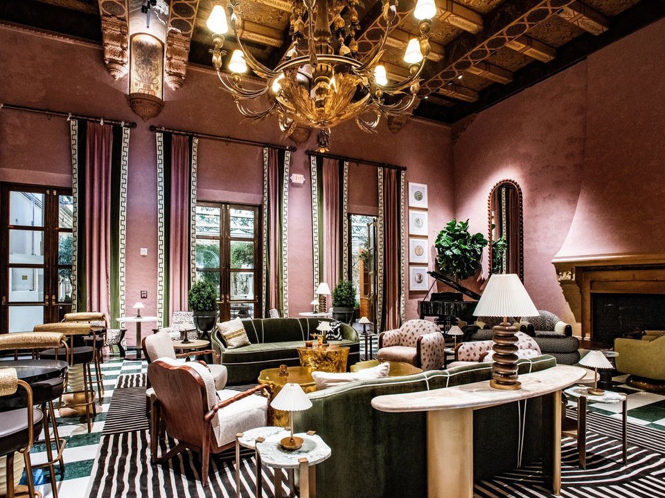 An elegant hotel lounge or restaurant interior with pink walls, ornate ceiling, grand chandelier, tall windows with decorative curtains, and mid-centu