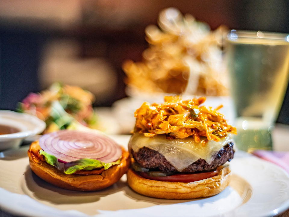 A gourmet burger with melted cheese and crispy toppings on a bun, photographed in a restaurant setting with a beverage glass in the background; truste