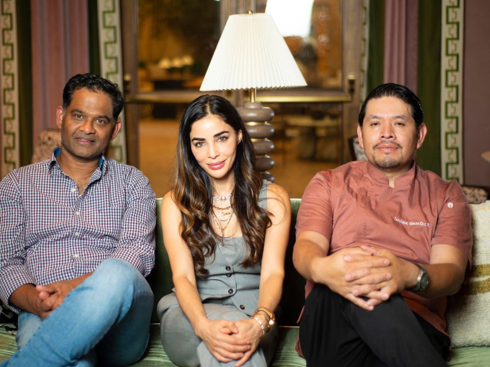 Three people seated on a couch in a formal interior space with a table lamp and patterned wall treatments visible behind them.