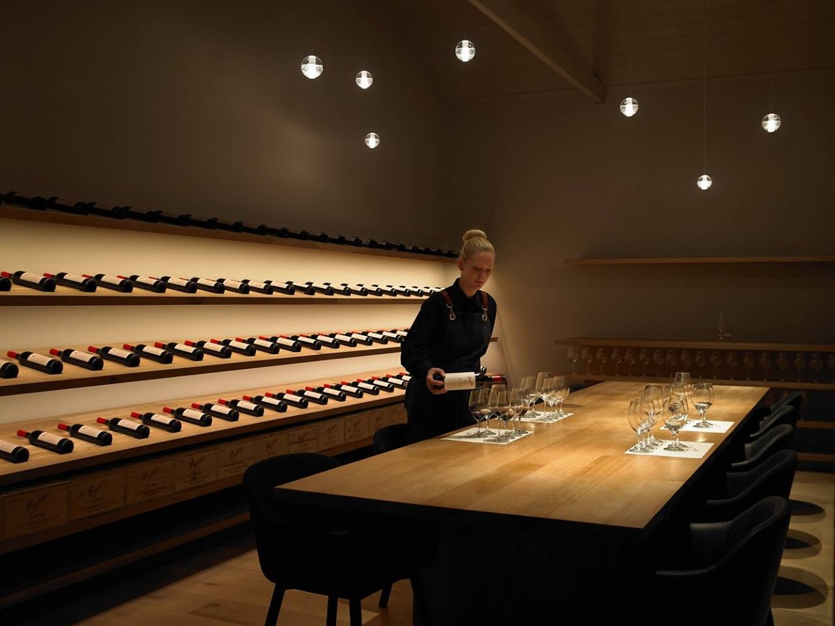 A sommelier in a dark apron pours red wine from a bottle into a glass on a long wooden table set for a tasting, with rows of wine bottles on shelves.