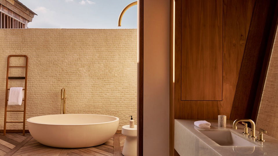 A luxury bathroom interior with a freestanding white tub, wooden deck flooring, beige mosaic tile wall, and a wooden vanity area with brass fixtures;